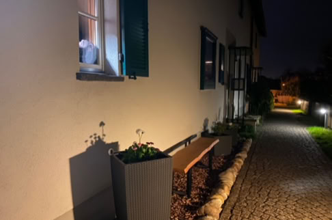 FHG am Hörnli, Außenansicht eines Hauses bei Nacht mit beleuchtetem Fenster, Bank, Pflanzkübeln und gepflastertem Weg.