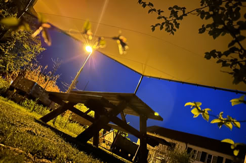 FHG am Hörnli, Holztisch unter einem Sonnensegel bei Nacht, mit Straßenlaterne im Hintergrund und umgebendem Laub.