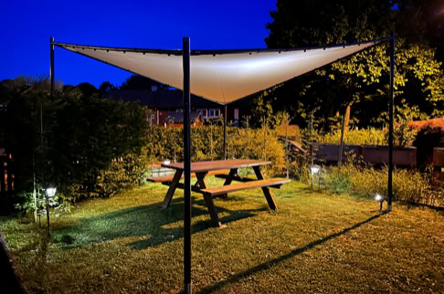 FHG am Hörnli, Ein Holztisch mit Bänken steht auf einer Wiese unter einem dreieckigen Sonnensegel, umgeben von Büschen und Bäumen bei Dämmerung.