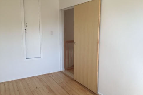 FHG am Hörnli, Leerer Raum mit Holzboden, weiß gestrichenen Wänden, einer Schiebetür und einer kleinen Wandöffnung.