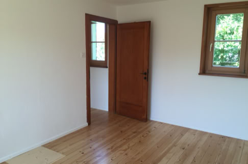 FHG am Hörnli, Leerer Raum mit Holzboden, einer Holztür und zwei Fenstern.