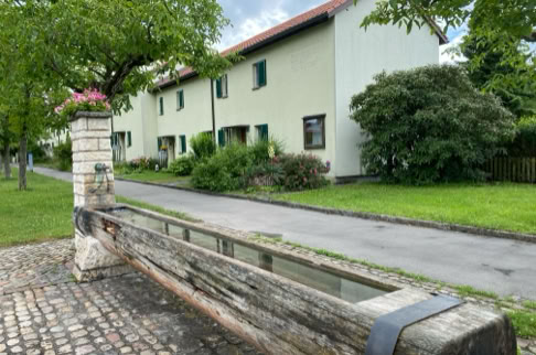 FHG am Hörnli, Holzbrunnen im Vordergrund, gepflasterter Boden, grüne Büsche und Bäume, zweistöckiges Gebäude im Hintergrund.