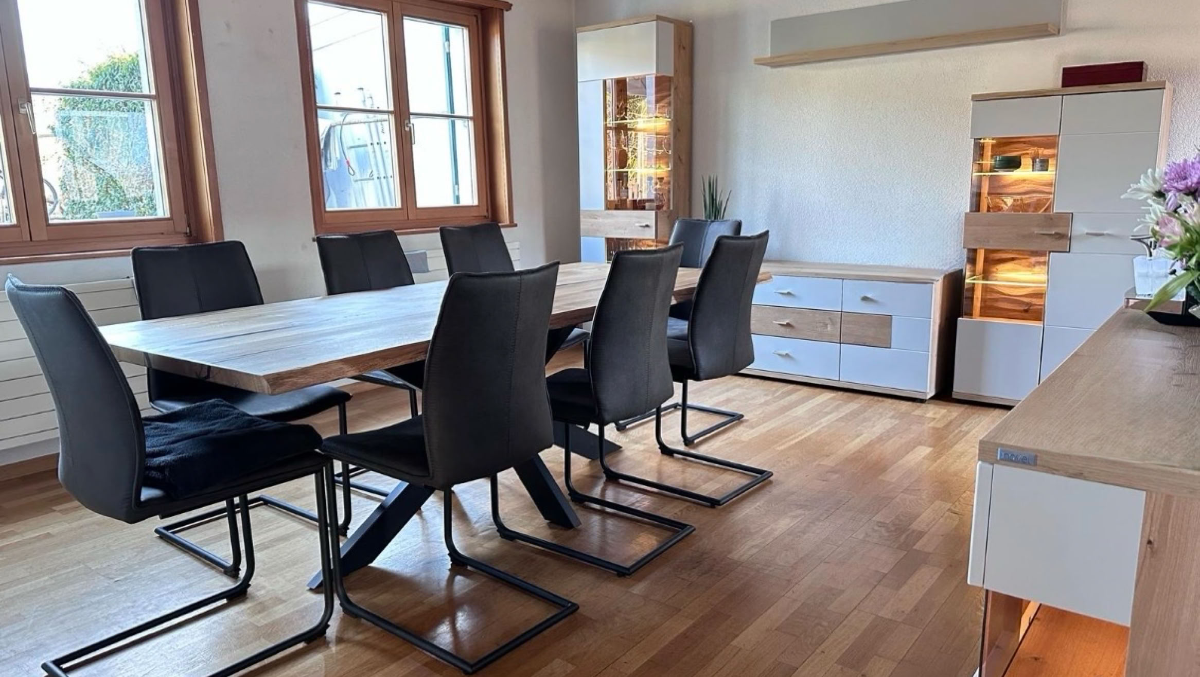 Esszimmer mit Holztisch und acht schwarzen Stühlen, Holzboden, zwei Fenstern, und einem beleuchteten Sideboard.