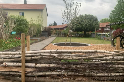 FHG am Hörnli, Garten mit einem Baum in der Mitte, umgeben von einem niedrigen Zaun aus Holzstämmen, im Hintergrund ein Haus mit rotem Dach und bewölktem Himmel.